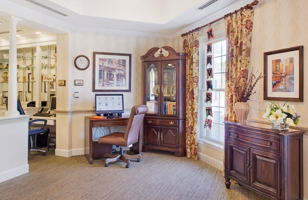 A traditional office interior featuring a wooden desk with a computer, an upholstered chair, and a large window with patterned curtains.