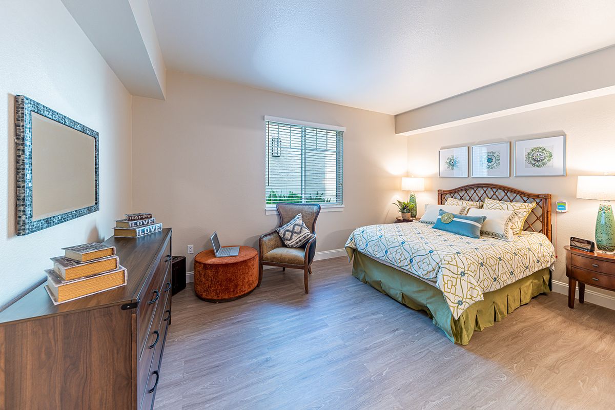 A cozy bedroom with a bed, chair, and window, featuring warm lighting and decorative elements.