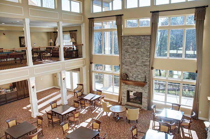 A spacious lounge area with high ceilings, large windows, and a stone fireplace, featuring multiple tables and chairs arranged for guests.
