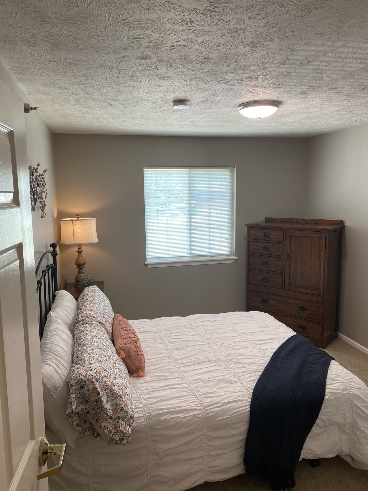 A cozy bedroom with a bed, dresser, and lamp near a window.