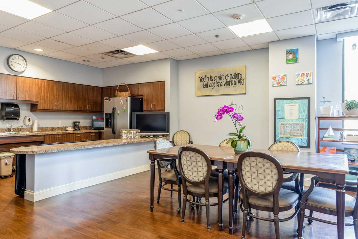 A communal kitchen and dining area featuring a table with chairs, a countertop with cabinets, and decorative flowers.