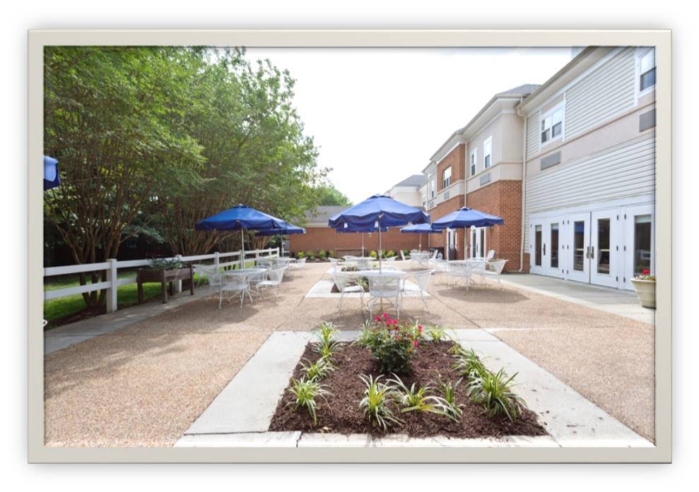 A well-maintained outdoor patio area with tables and blue umbrellas, surrounded by landscaping and adjacent to a building.