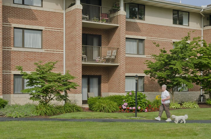 A senior person walks along a pathway in front of a brick apartment building with well-maintained landscaping.