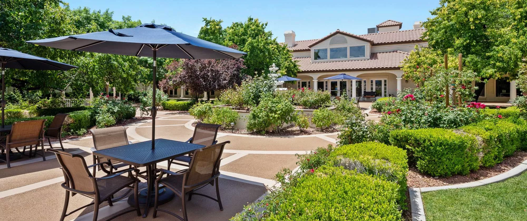 A serene garden with outdoor seating and umbrellas, surrounded by lush greenery on a sunny day.
