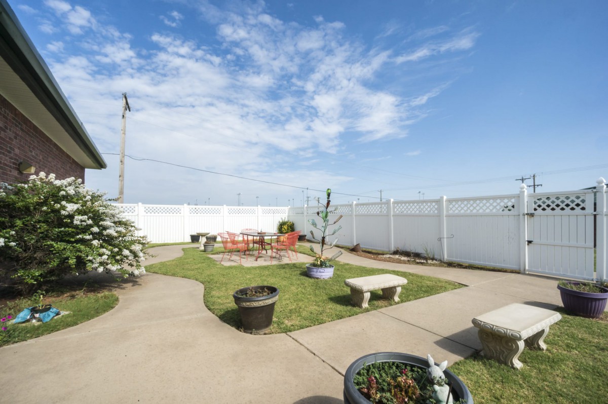 A sunny backyard garden with a patio, benches, and plants surrounded by a white fence.