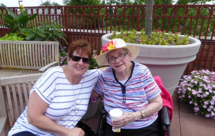 Two women sitting outside, smiling, with one in a wheelchair wearing a floral hat.