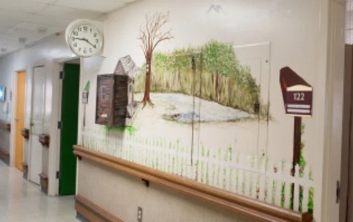 A hospital hallway features a mural depicting a serene nature scene with a tree and pond, accompanied by a clock on the wall.