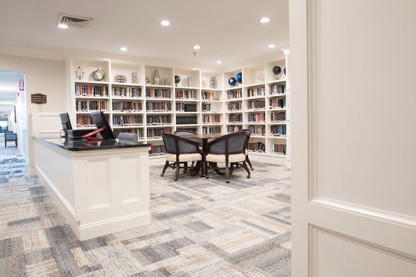 A cozy library with white bookshelves, a central table surrounded by chairs, and bright overhead lighting.
