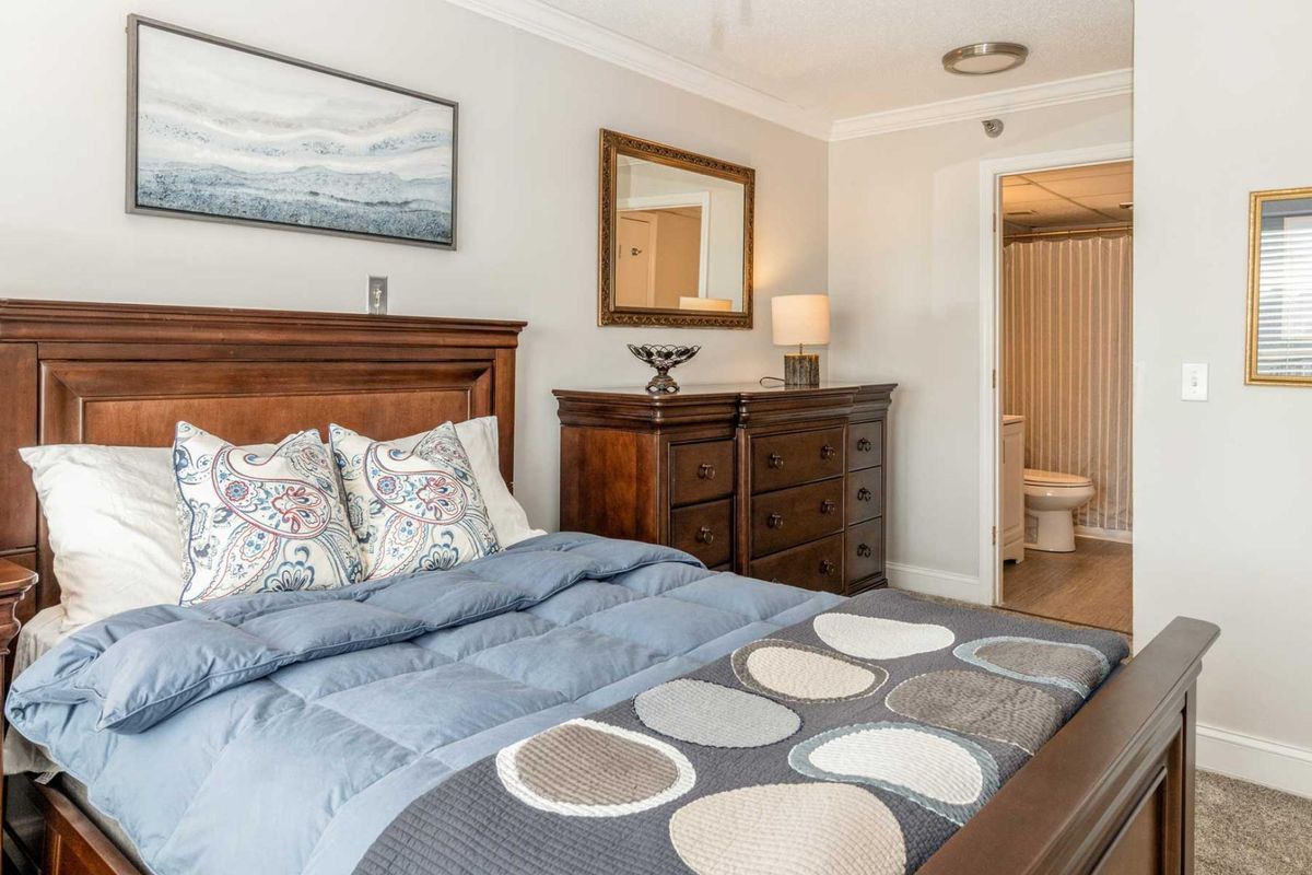 A cozy bedroom with a neatly made bed, wooden dresser, mirror, and decorative lamp.