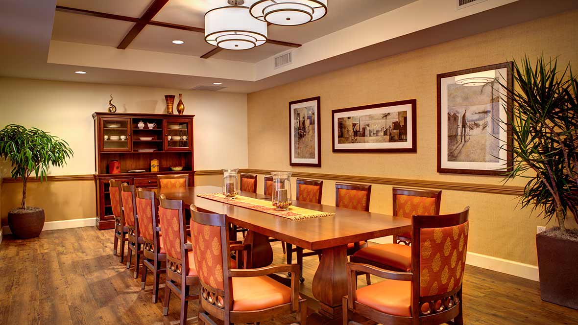 A modern dining room with a long wooden table and orange cushioned chairs, featuring elegant lighting and framed wall art.