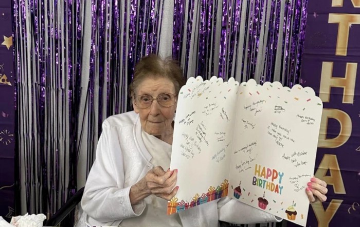 An elderly person holding a large birthday card with messages written inside, surrounded by purple decorations.