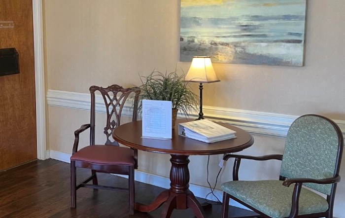 A cozy corner with a round wooden table, two chairs, a table lamp, and wall art.