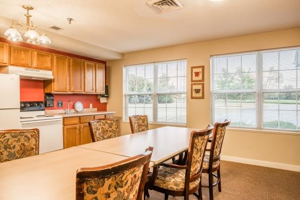 A cozy dining area with a wooden table and patterned chairs, adjacent to a kitchen with warm wooden cabinetry and large windows.