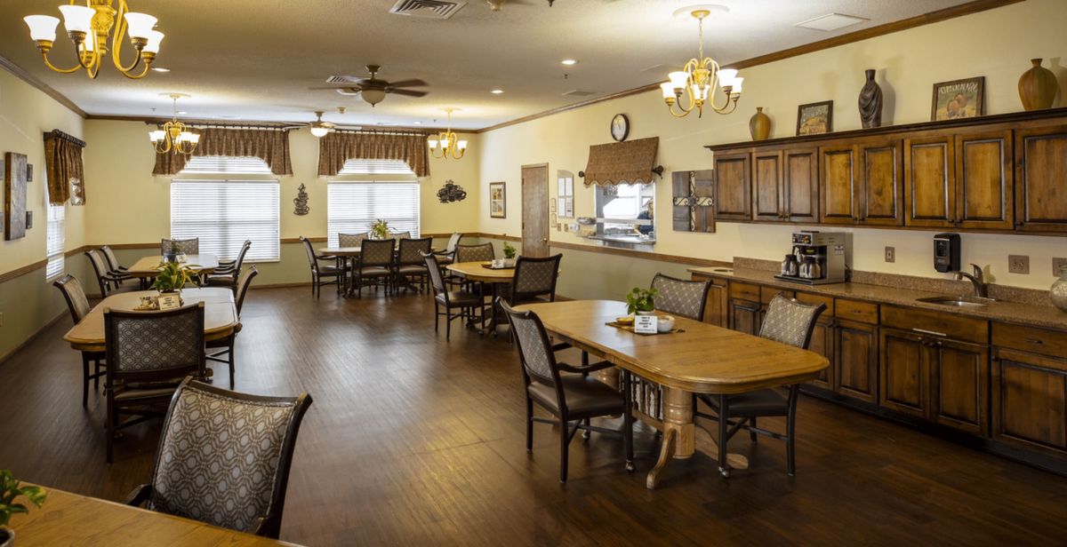 A spacious dining room with wooden tables and chairs, featuring a kitchen area and elegant chandeliers.