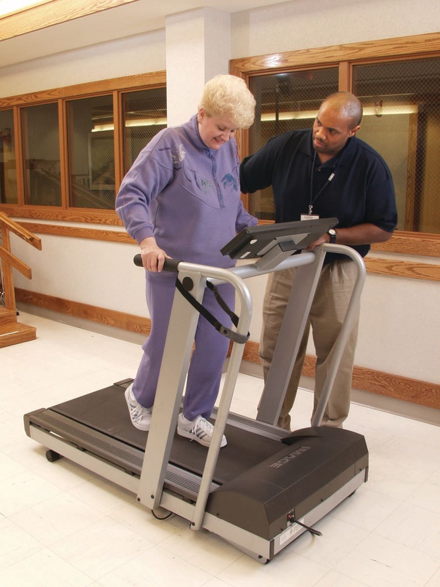 An elderly woman receiving assistance while walking on a treadmill for physical therapy.