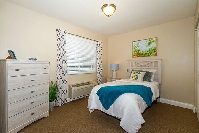 A cozy bedroom with a neatly made bed, a lamp on the bedside table, and a dresser near the window.