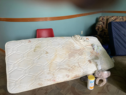 A stained mattress laid out in a room with a red chair and cleaning supplies nearby.
