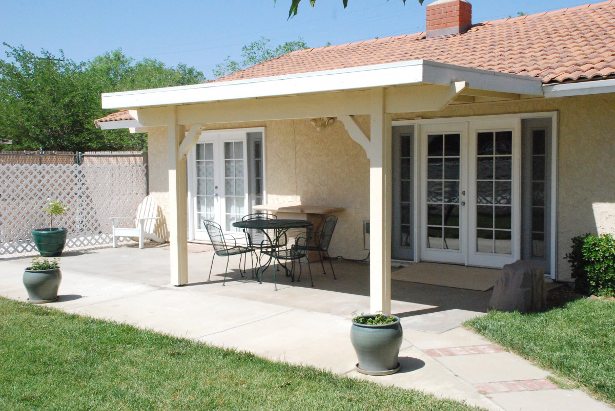 A cozy patio area with a table and chairs under an awning, set against a house with glass doors leading to a backyard.