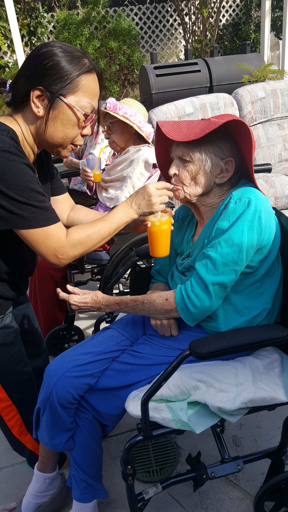 A caregiver assists an elderly woman in a wheelchair by holding a drink for her outdoors.