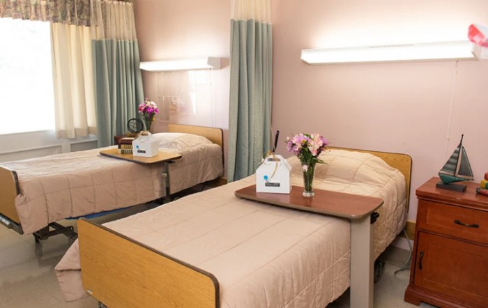 A hospital room with two neatly made twin beds, equipped with tray tables and adorned with flower arrangements.
