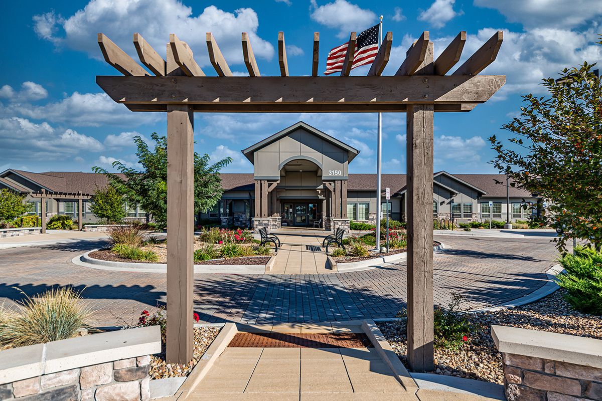 Elegant building entrance with a pergola and American flag under a blue, cloudy sky.
