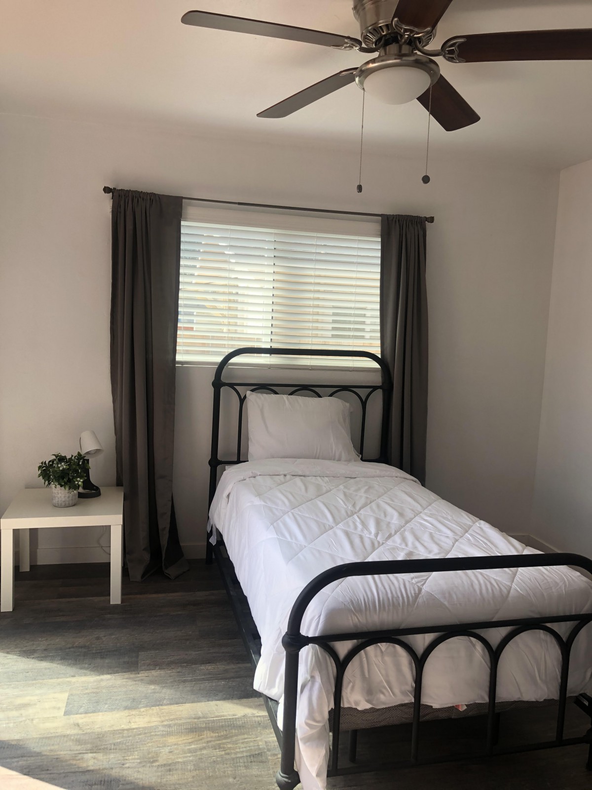 A minimalist bedroom with a single metal-framed bed and a nightstand by the window, accented by dark curtains.