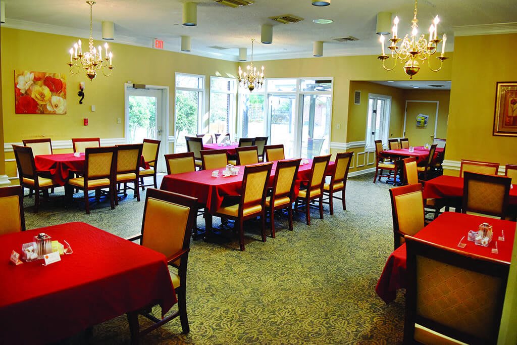 A spacious dining room with neatly arranged tables and red tablecloths, illuminated by elegant chandeliers.