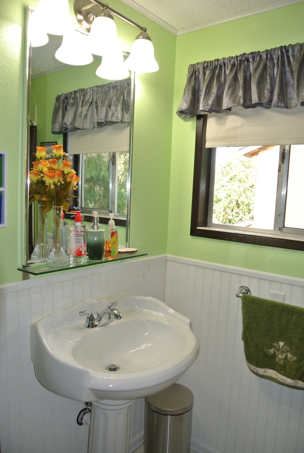 A cozy bathroom with green walls, featuring a sink, mirror, and window adorned with flowers and decor.