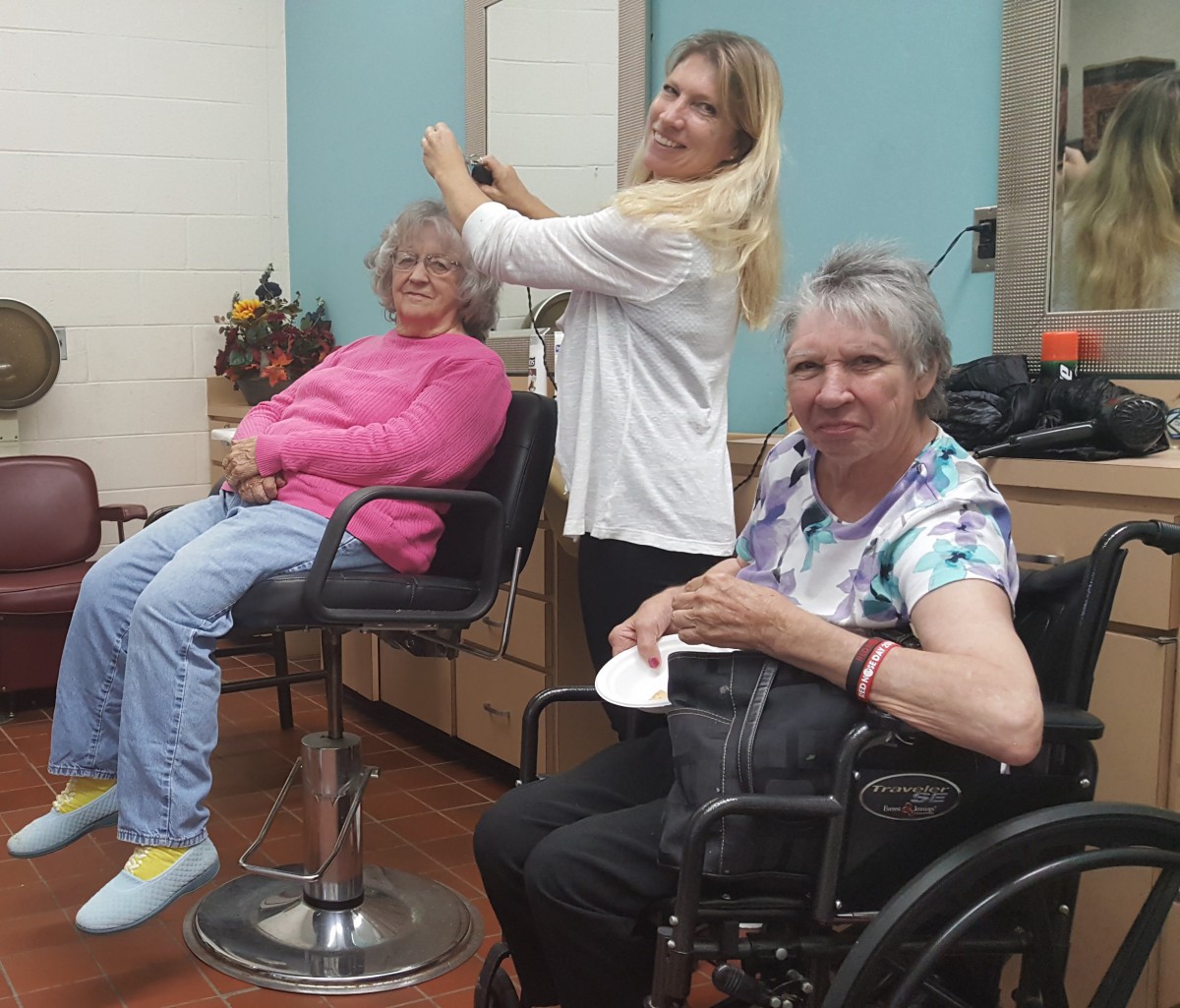 A hairdresser styling the hair of an elderly woman while another woman in a wheelchair smiles at the camera.