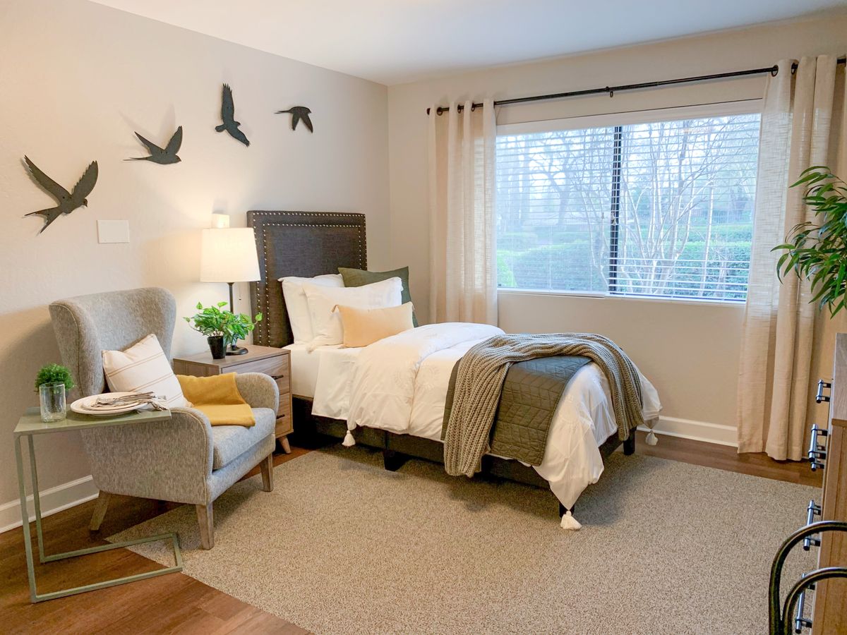 A cozy bedroom interior with a neatly made bed, a comfortable chair beside a lamp, and wall decorations of birds in flight.