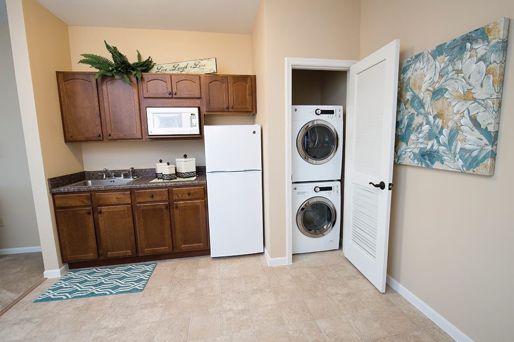 A compact kitchen with wooden cabinets, a microwave, and a refrigerator, featuring a stacked washer and dryer in an open closet.