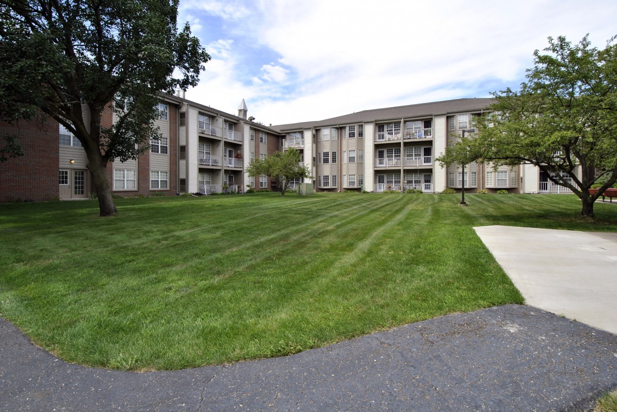 A three-story apartment building with a well-maintained lawn and trees under a partly cloudy sky.