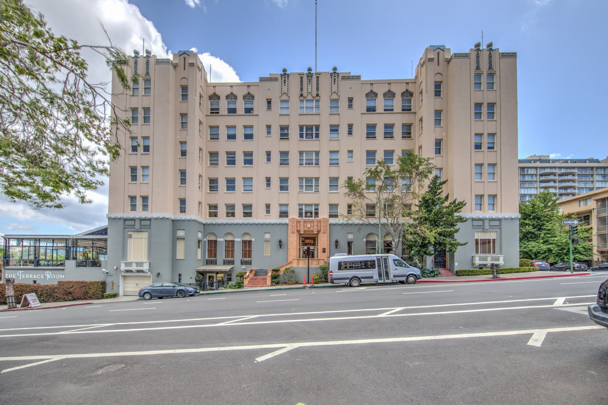 A large urban building with a street view, featuring several vehicles parked in front and trees lining the sidewalk under a partly cloudy sky.