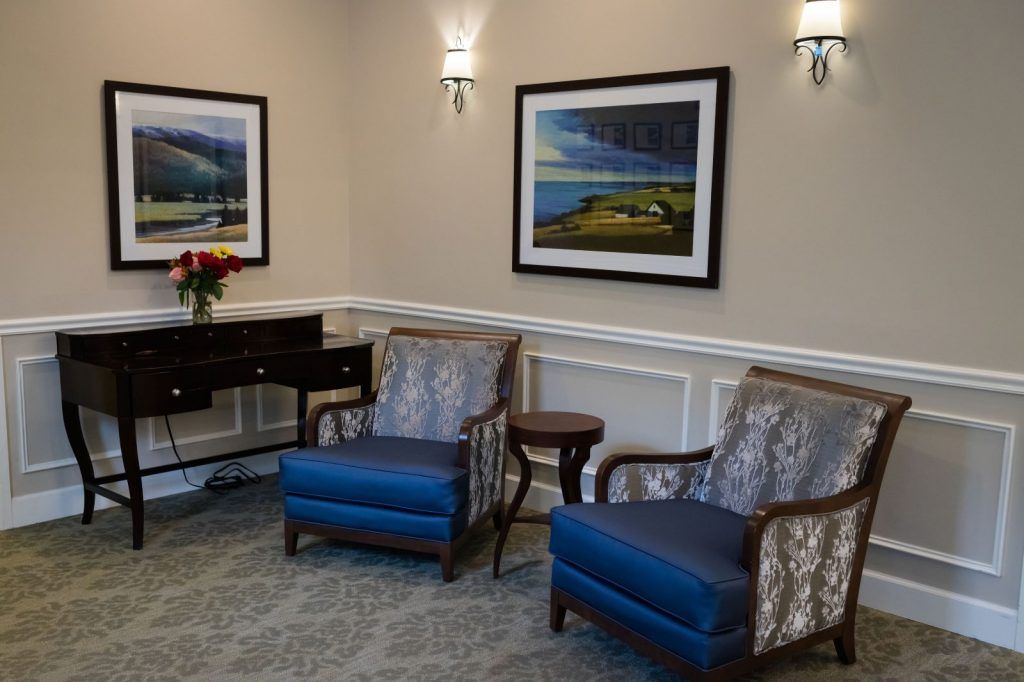 A cozy waiting room featuring two patterned chairs with blue cushions, artwork on the walls, a small table, and decorative lighting.