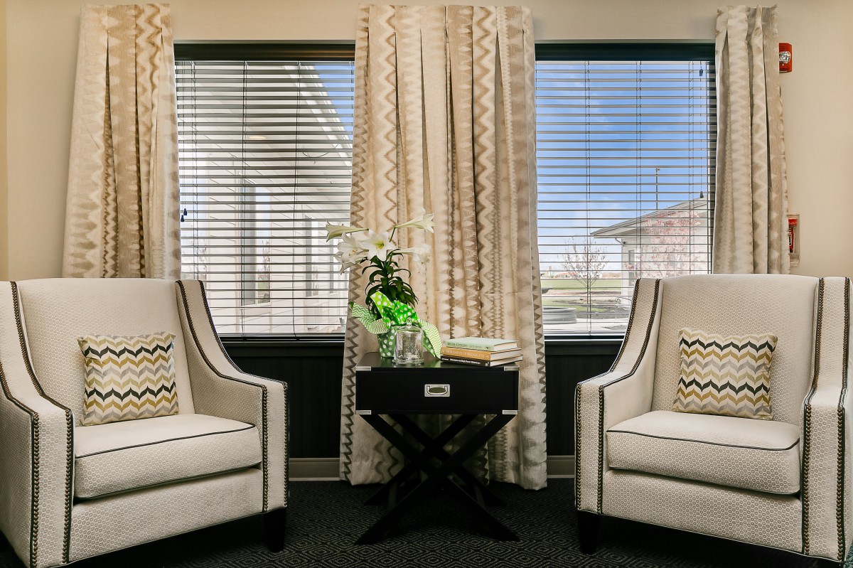 A cozy living room setup with two armchairs and a small table between them, adorned with a plant, framed by large windows with patterned curtains.