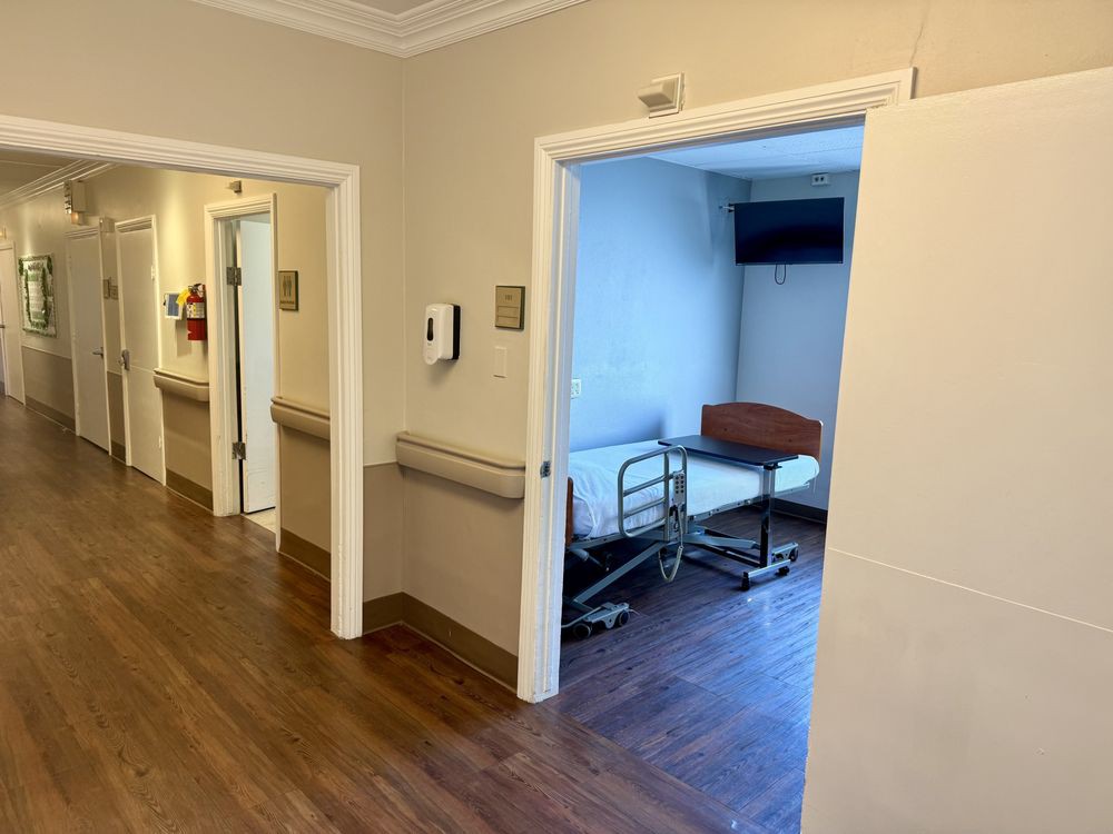 A hospital corridor with an open door leading to a patient room equipped with a bed.