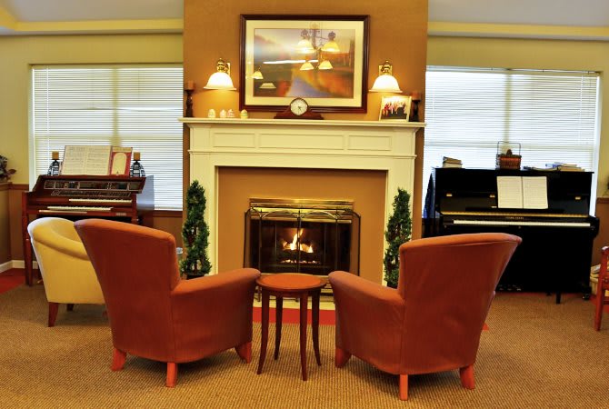 A cozy living room with two chairs facing a lit fireplace, flanked by lamps and decorated with artwork.