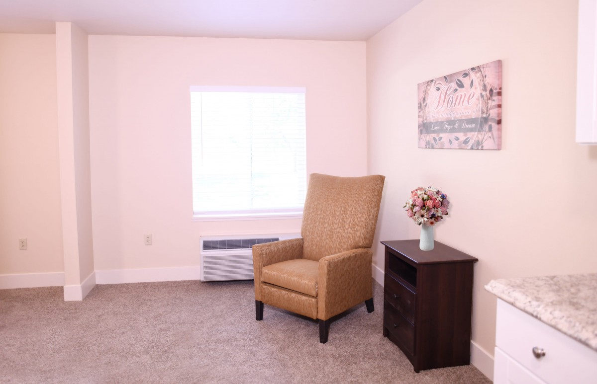 A minimalist room featuring a comfortable armchair, a small cabinet with a floral arrangement, and a large window providing natural light.