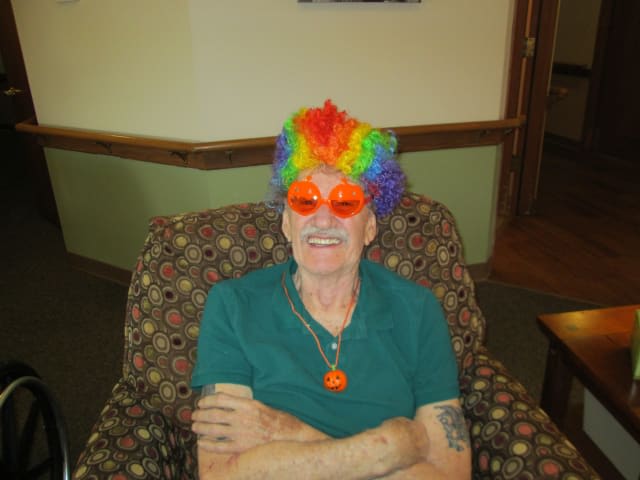 A person wearing a rainbow wig and orange sunglasses, sitting and smiling indoors.