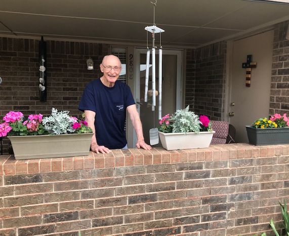 A man smiling while standing on a porch with flowers and a wind chime, leaning on a brick wall.