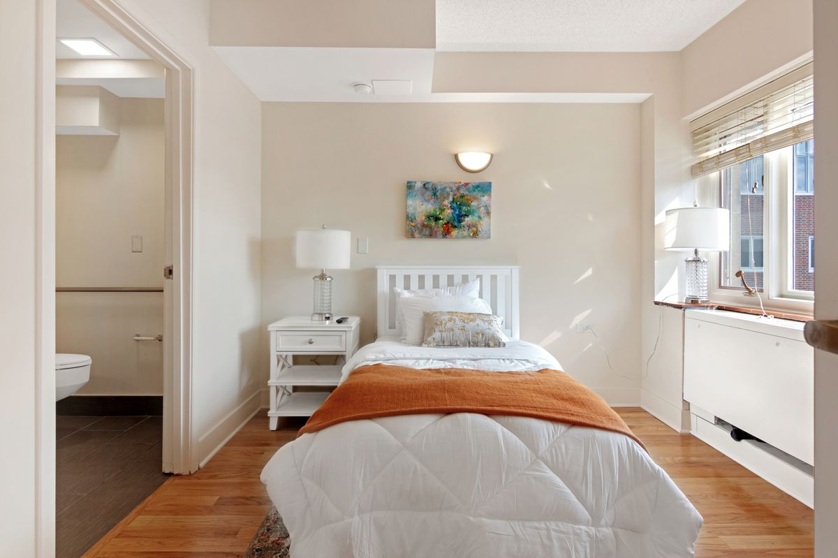 A cozy bedroom features a single bed with an orange blanket, complemented by a nightstand and lamp. Artwork adorns the wall above the bed, and sunlight streams through a window onto the hardwood floor.