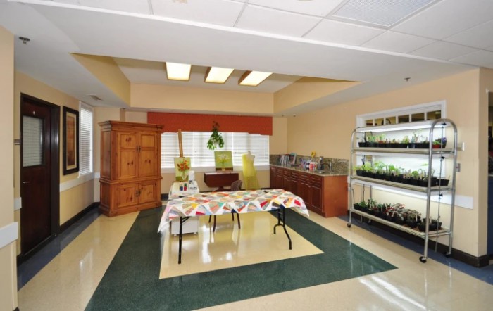 An indoor room featuring a table covered with a colorful cloth, surrounded by plants and shelves with additional greenery.