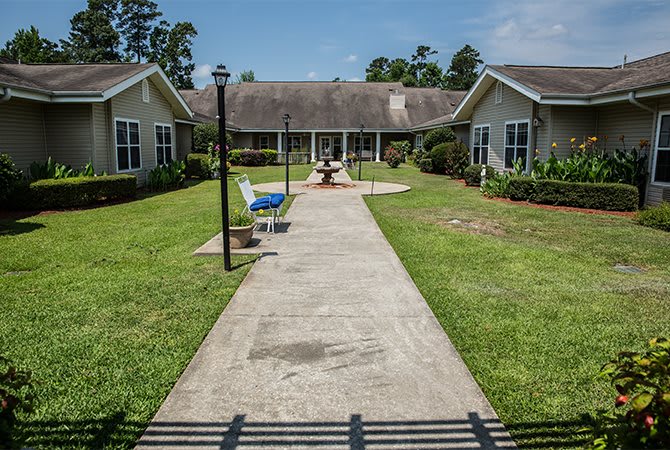 A pathway leading through a neatly landscaped area between two residential-style buildings, featuring benches and a well-maintained lawn.