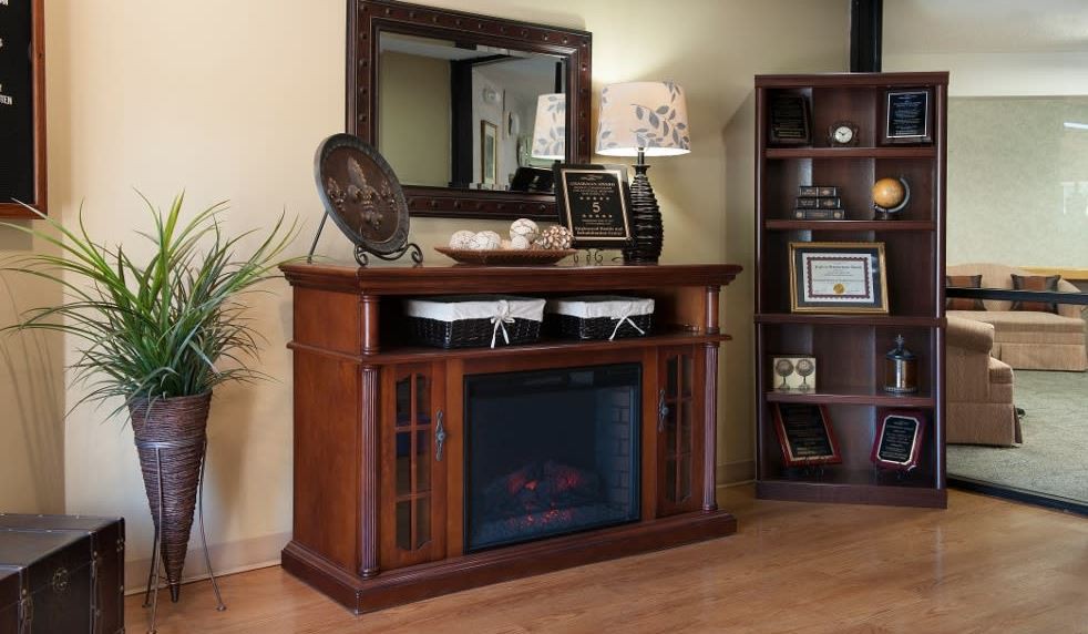 A cozy living room setup featuring a wooden fireplace console with decorative items, a large mirror above, and a bookshelf on the side.
