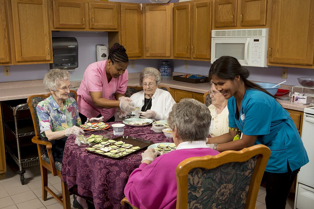 Caregivers assisting elderly women during a meal in a kitchen setting.