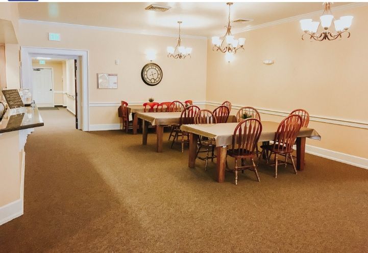 A spacious dining room with wooden tables and chairs, illuminated by chandeliers, featuring a carpeted floor and a wall-mounted clock.