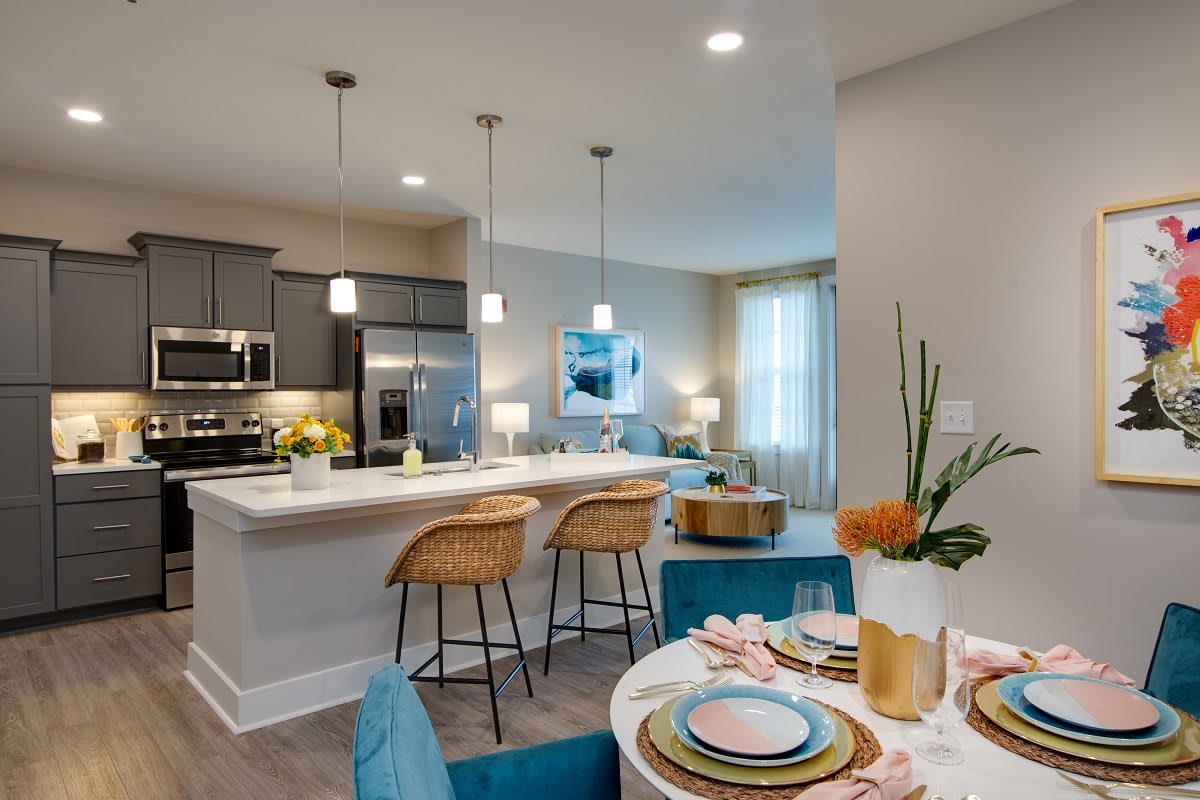 A modern kitchen and dining area featuring sleek design with pendant lighting, a breakfast bar with woven stools, and a stylishly set dining table.