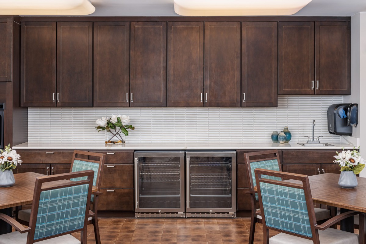 A modern kitchen with dark wooden cabinets, a sleek countertop, and dual wine fridges, accented by decorative flowers and chairs.