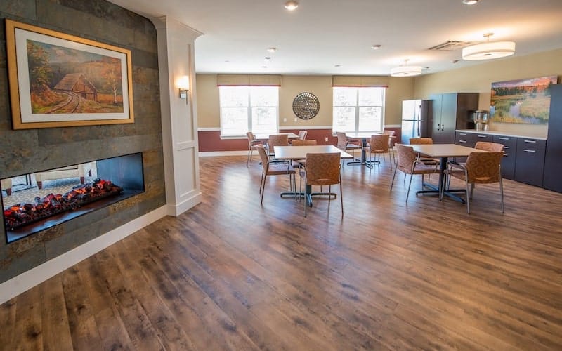 A cozy dining area with hardwood floors, tables, and chairs, illuminated by natural light from large windows.