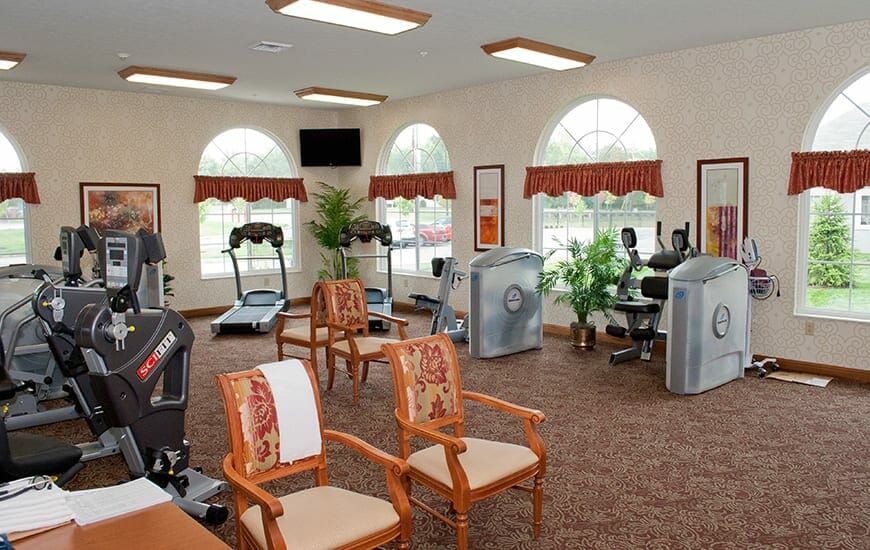 A well-lit exercise room featuring cardio equipment, chairs, and indoor plants.
